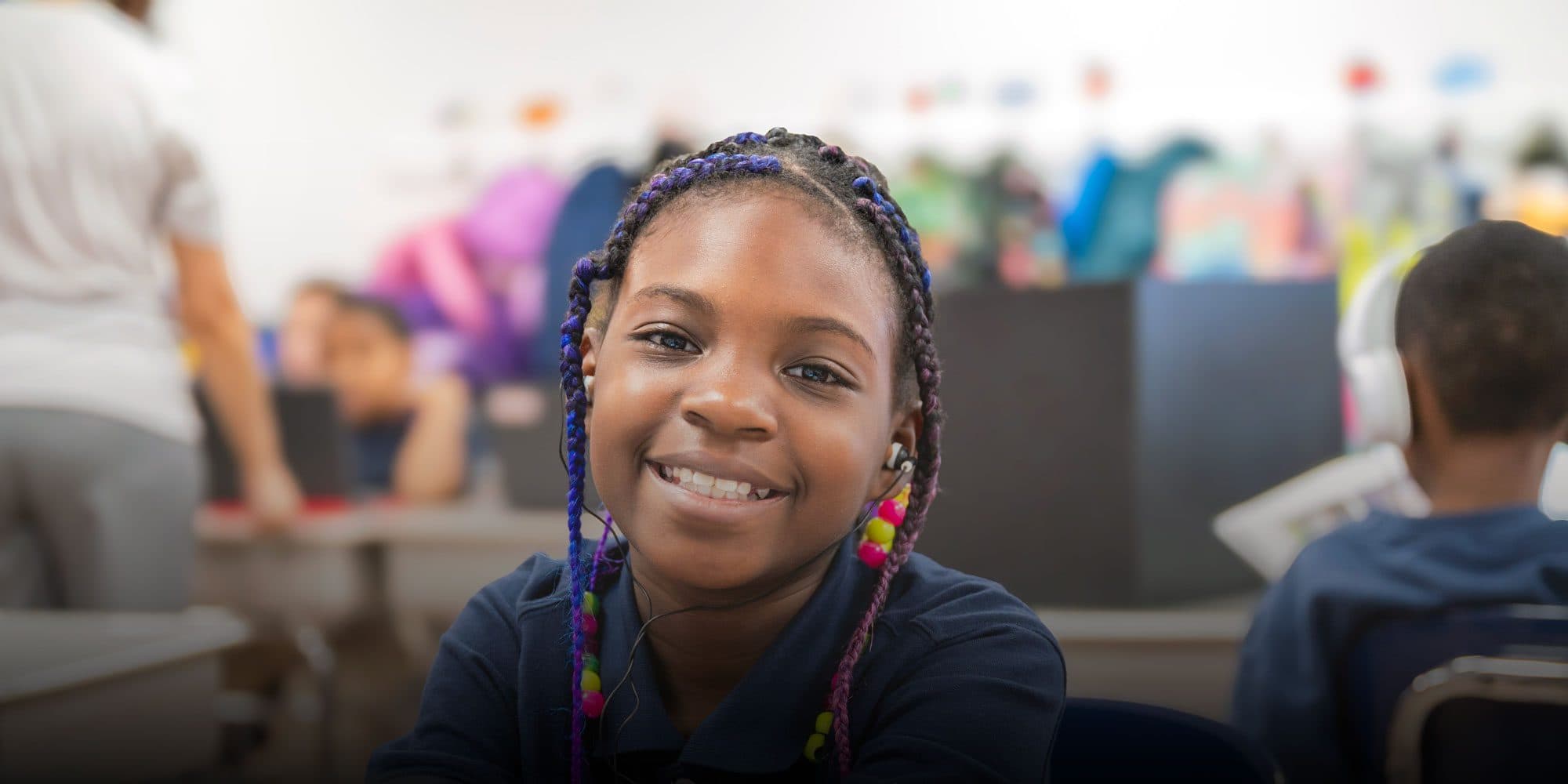 Smiling student with braided hair and earbuds in a classroom.