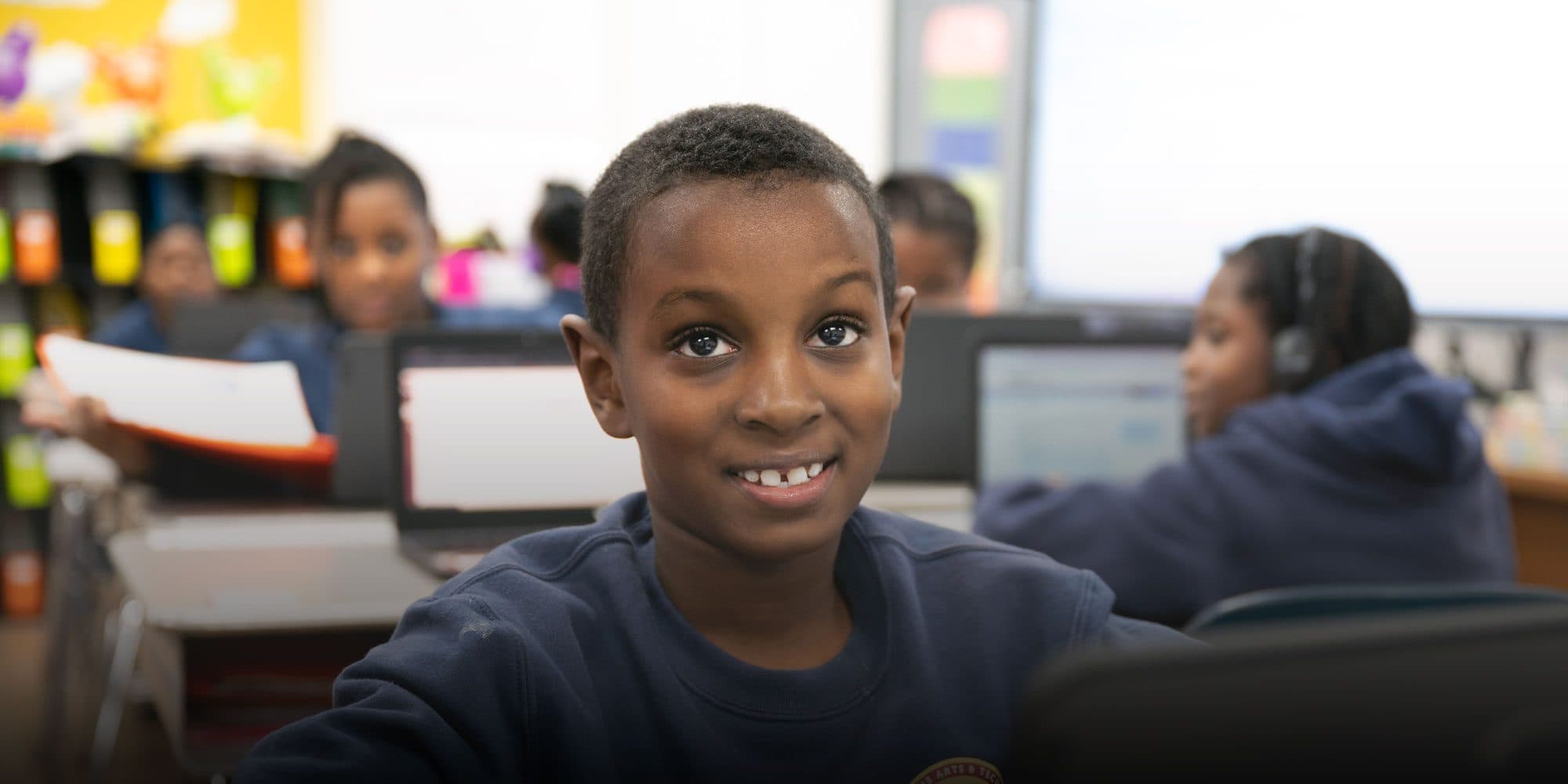 Smiling student at a desk in a classroom with laptops.