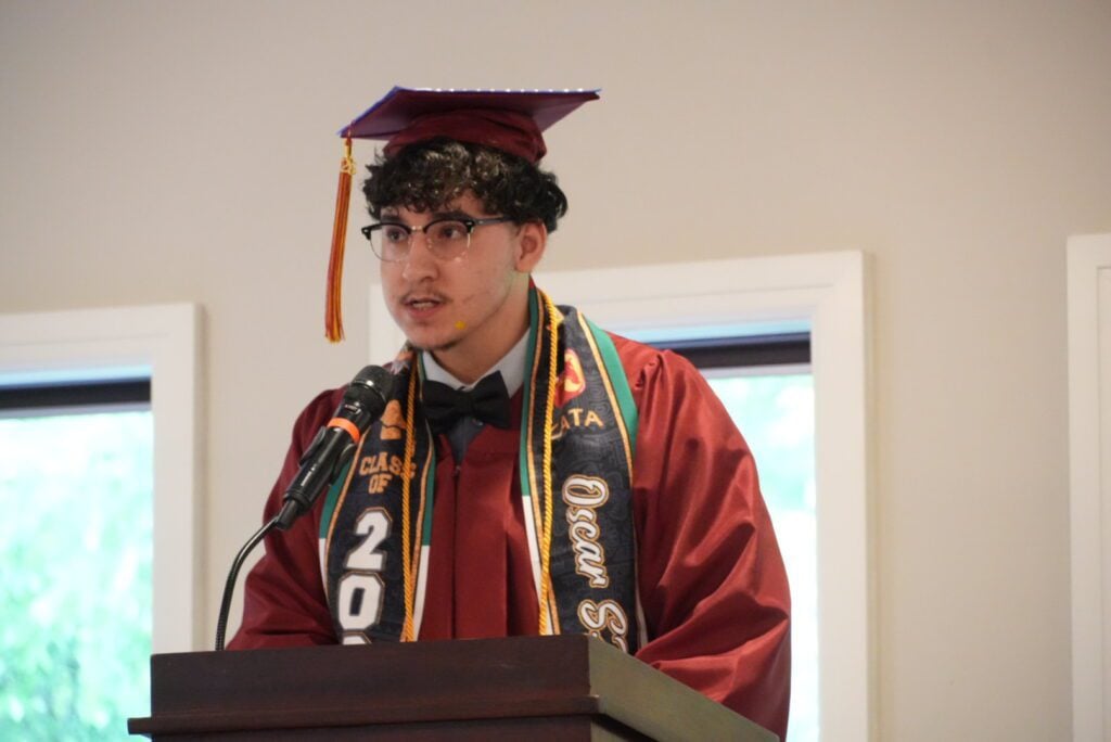 Adult in graduation cap and gown speaking at podium.