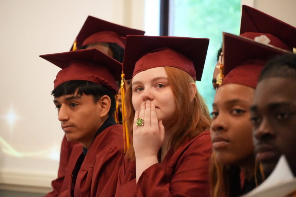 Students in maroon graduation caps and gowns seated, one with hand over mouth.