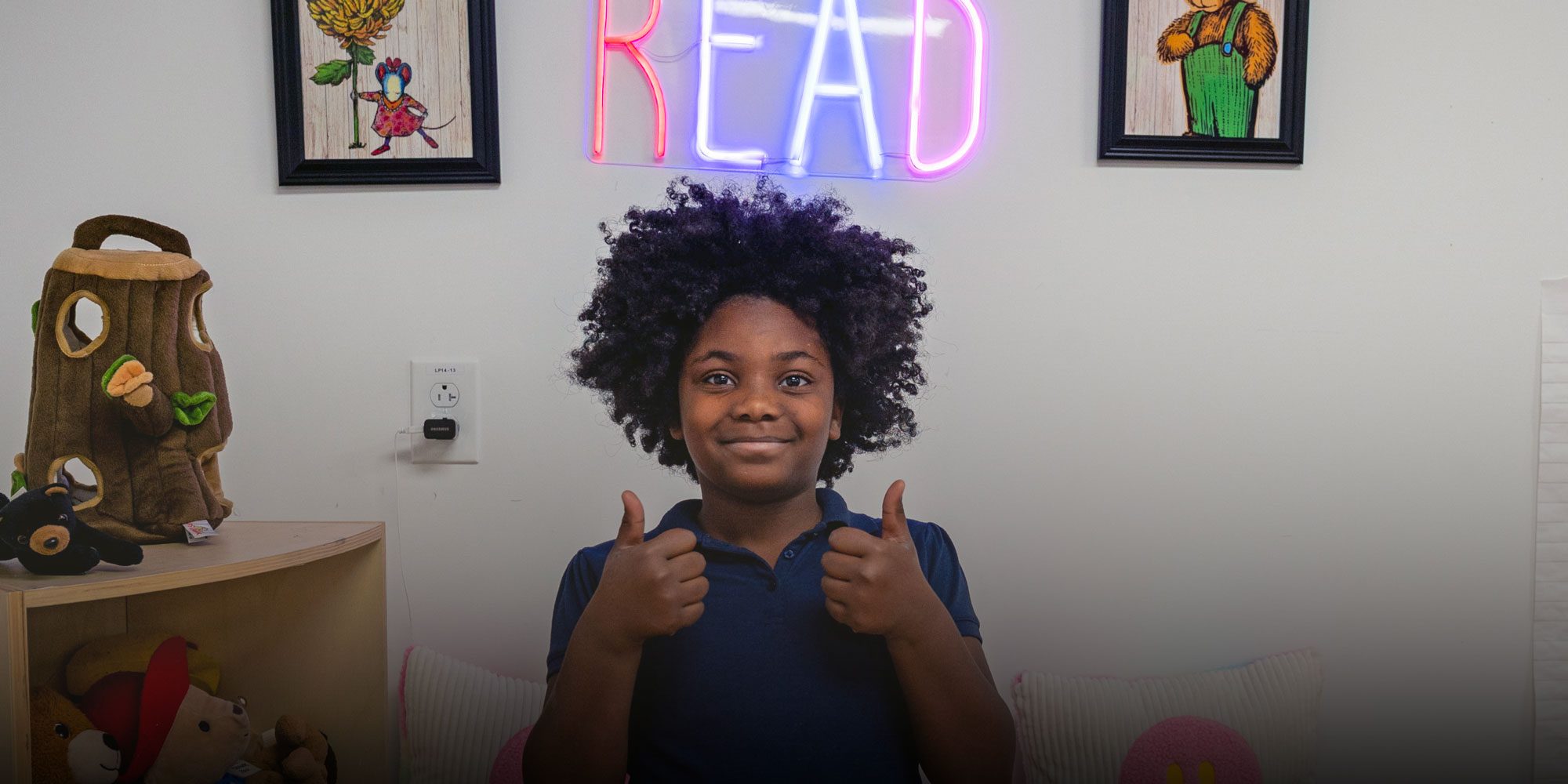 Smiling student giving two thumbs up in a reading nook under a neon READ sign.